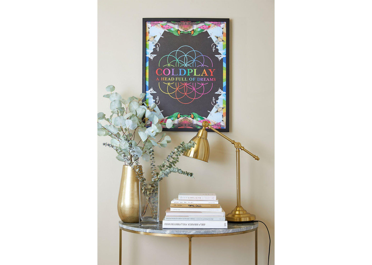 A Head Full Of Dreams Lithograph, hung above a small table shaped like a crescent moon, with a marble top and golden legs. The table has golden and clear vases with eucalyptus, books and a golden desk lamp. The poster is on a black wooden frame. The colorful artwork contrasts with the beige wall